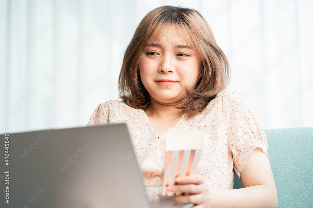 Young Asian girl sitting on sofa eating and watching movies on laptop