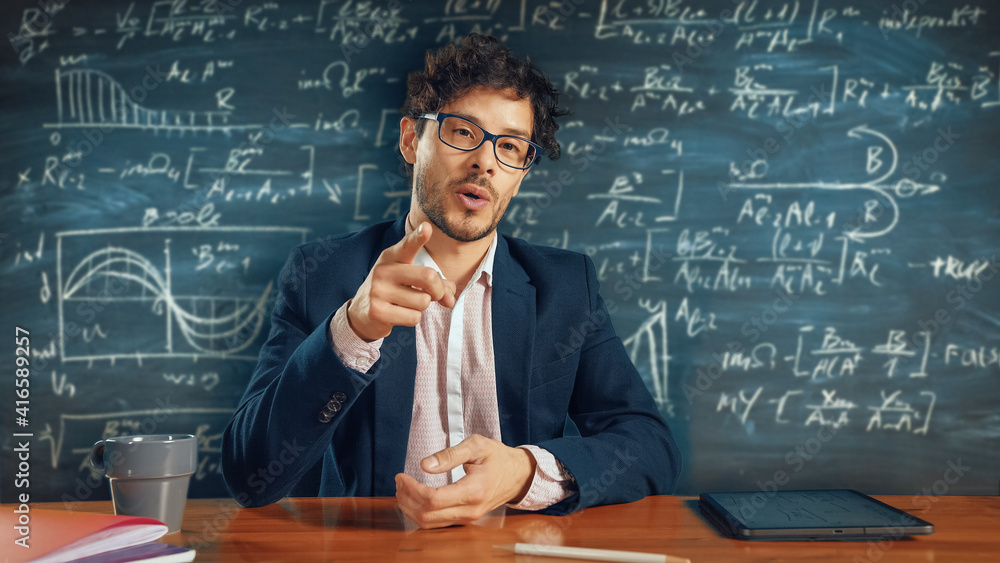 Online e-Education: Teacher Sitting at Desk, Explains Lesson to a Classroom, Behind Him Blackboard w