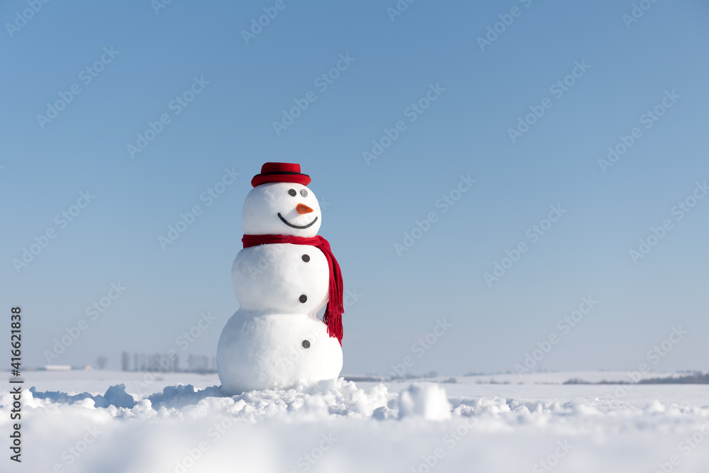 Funny snowman in stylish red hat and red scalf on snowy field. Blue sky on background