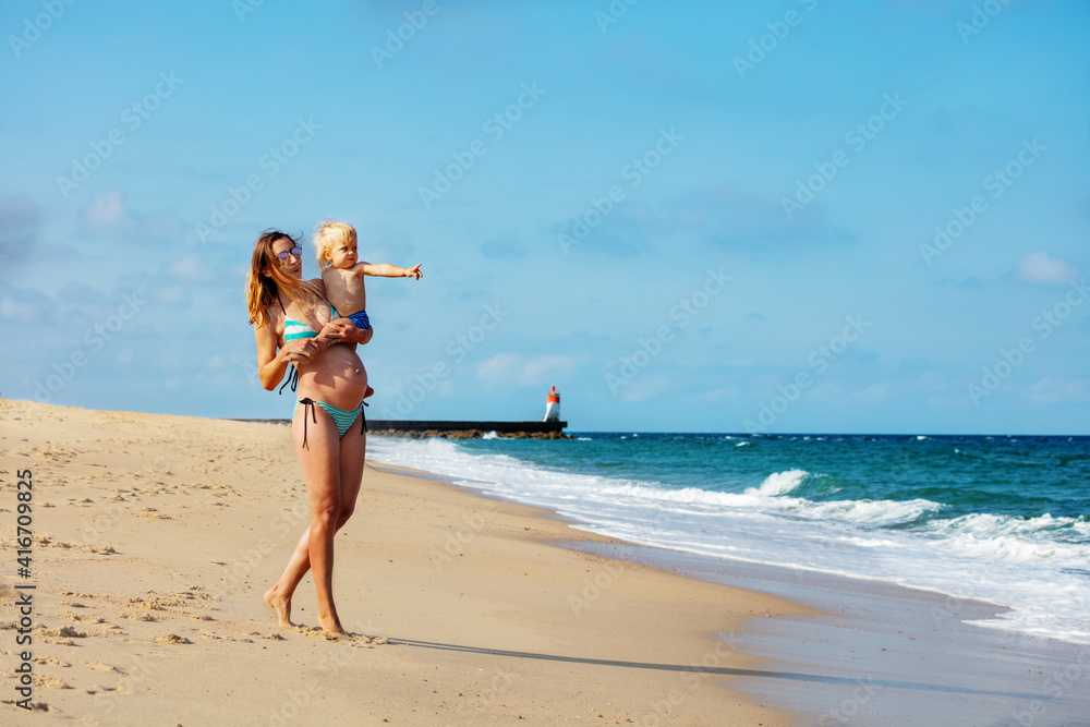 Happy pregnant mother hold toddler boy point with finger in hand and standing on the sand sea beach 