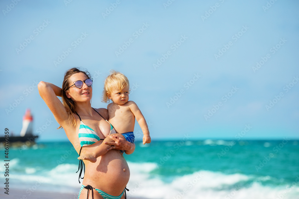 孕妇抱着金发蹒跚学步的孩子站在海边，背后有灯塔的近照