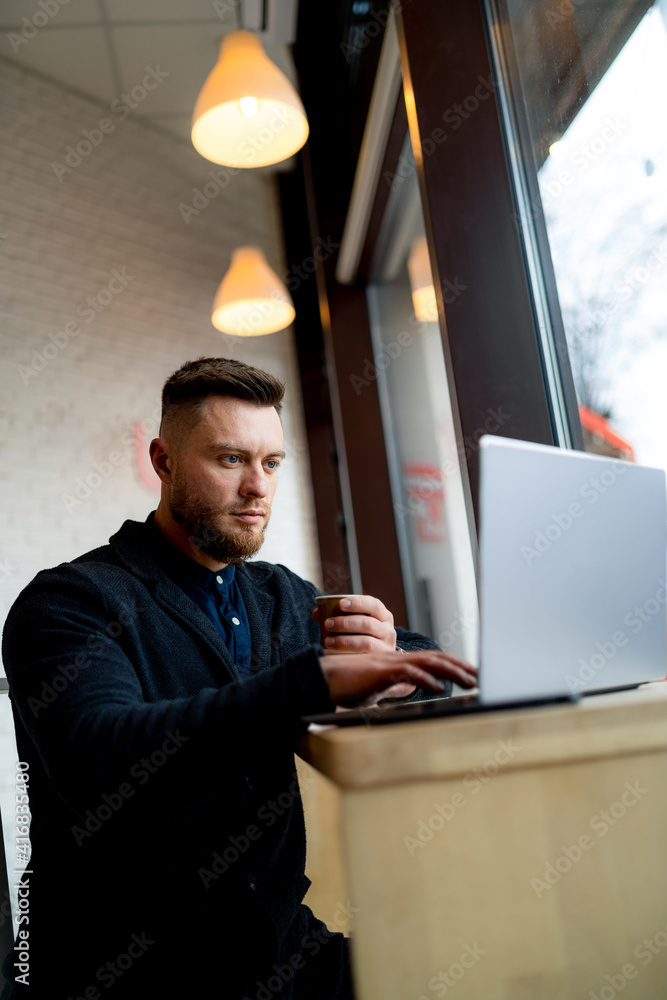 Bearded fashion young manager works on laptop while having coffee break. Modern interior of cafe. Cl