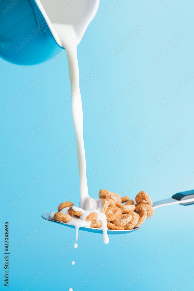 Pouring of milk from jug into spoon with cereal rings on color background