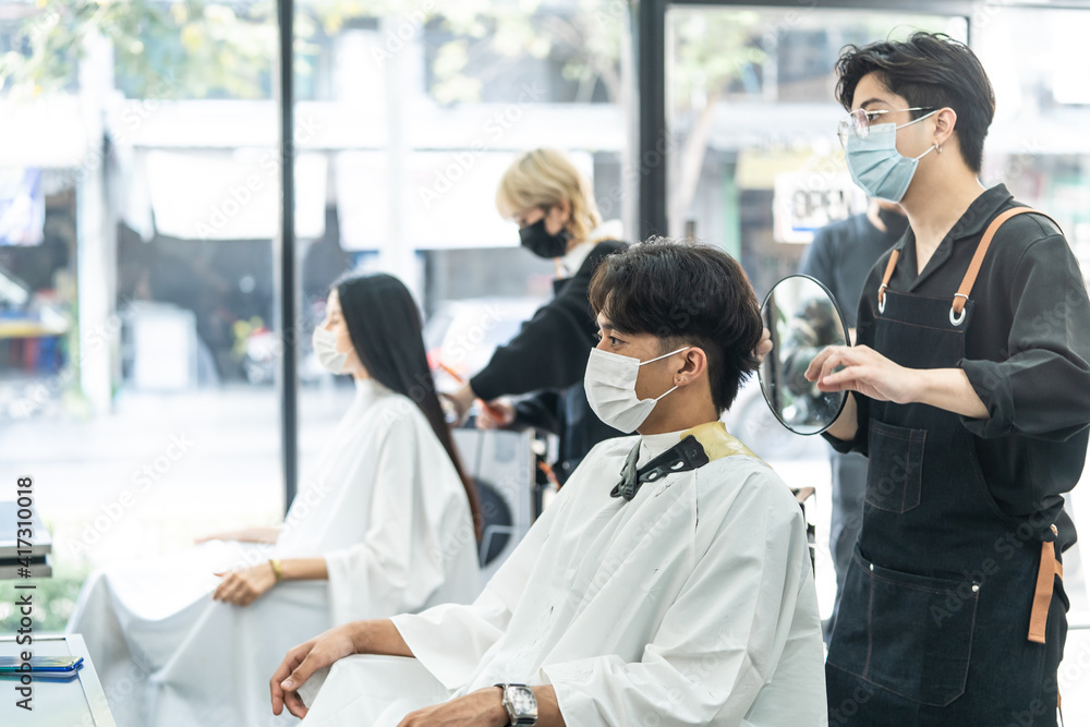 Professional male stylist cutting mans hair in salon. The man wearing mask and face shield to preve