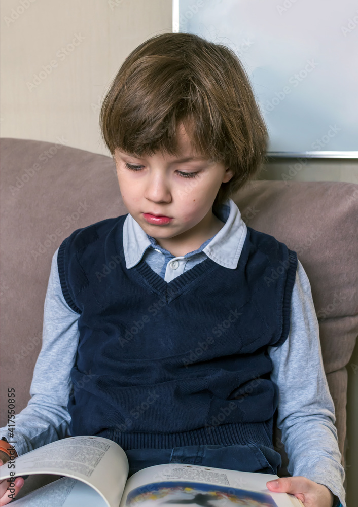 An caucasian eight-year-old boy in a school uniform is sitting on the couch and reading a book. Port