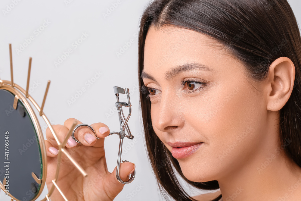 Beautiful young woman with eyelash curler and mirror against light background