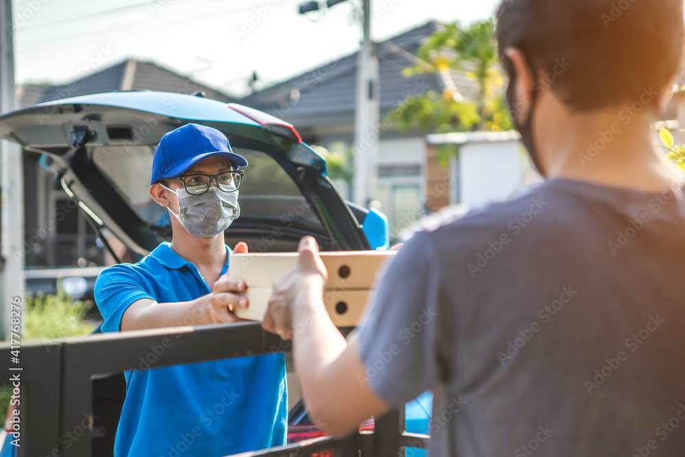 Asian delivery man wearing mask delivers pizza, customer in medical gloves signs on tablet. Delivery