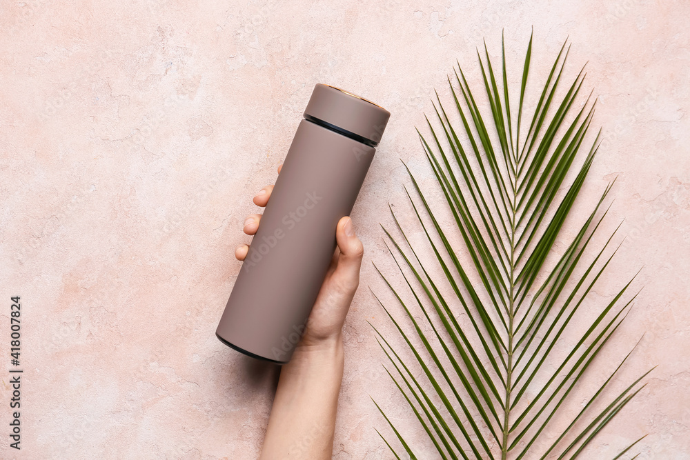 Female hand with modern thermos and tropical leaf on color background