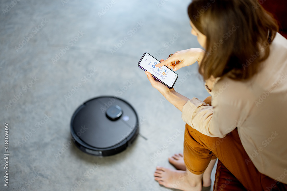 Black robotic vacuumer is cleaning the floor while pretty woman control the robot with Smartphone an