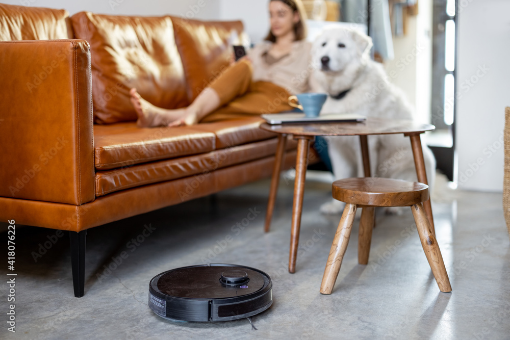 Black robotic vacuumer is cleaning the floor while pretty woman sitting on the brown sofa with a whi