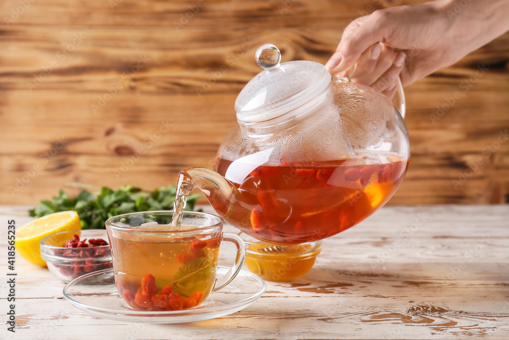 Woman pouring goji tea from teapot into cup on wooden background