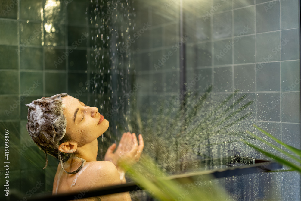 Woman staying back in shower apply shampoo with water dripping beside the glass wall. Taking shower 