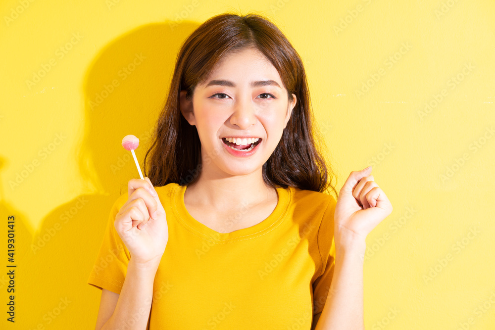 Young Asian woman eating lollipop on yellow background