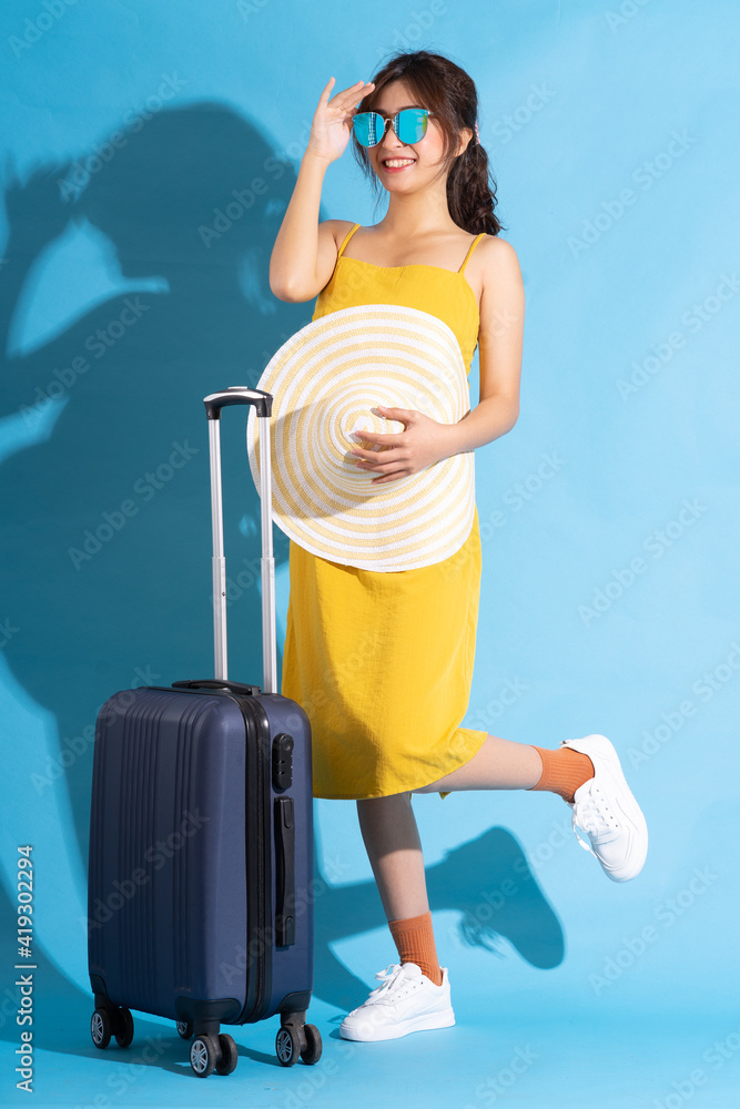 Young Asian woman holding suitcase on blue background, summer concept