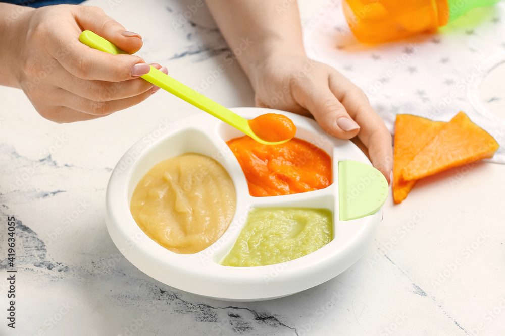 Woman holding plate with different baby food on light background, closeup