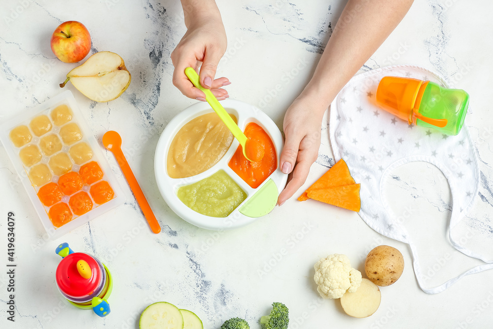 Woman holding plate with different baby food on light background