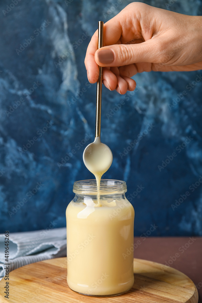 Female hand, spoon and jar with sweet condensed milk on color background