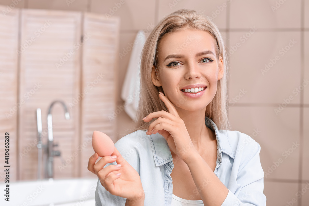 Beautiful young woman with sponge applying makeup at home