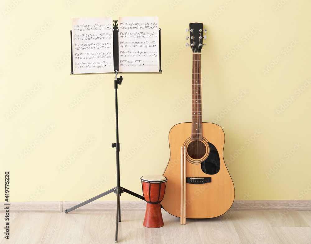 Note stand with music sheets, acoustic guitar, djembe and drumsticks near color wall