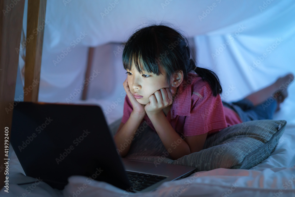 Asian girl child to make a camp to play imaginatively watching a film on laptop in the darkness of t