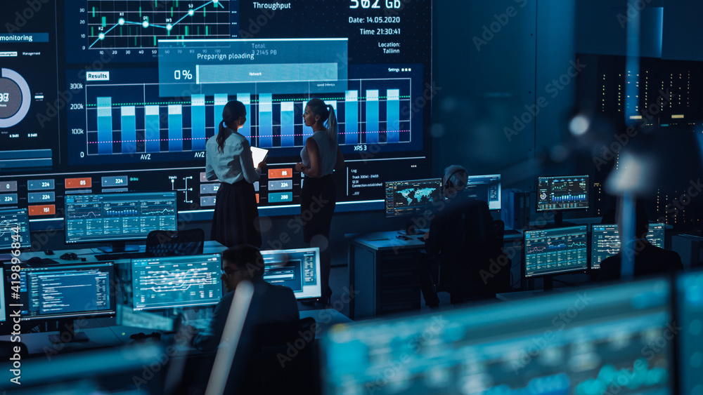 Two Software Engineers Talking Next to a Big Screen in a Modern Monitoring Office with Live Analysis