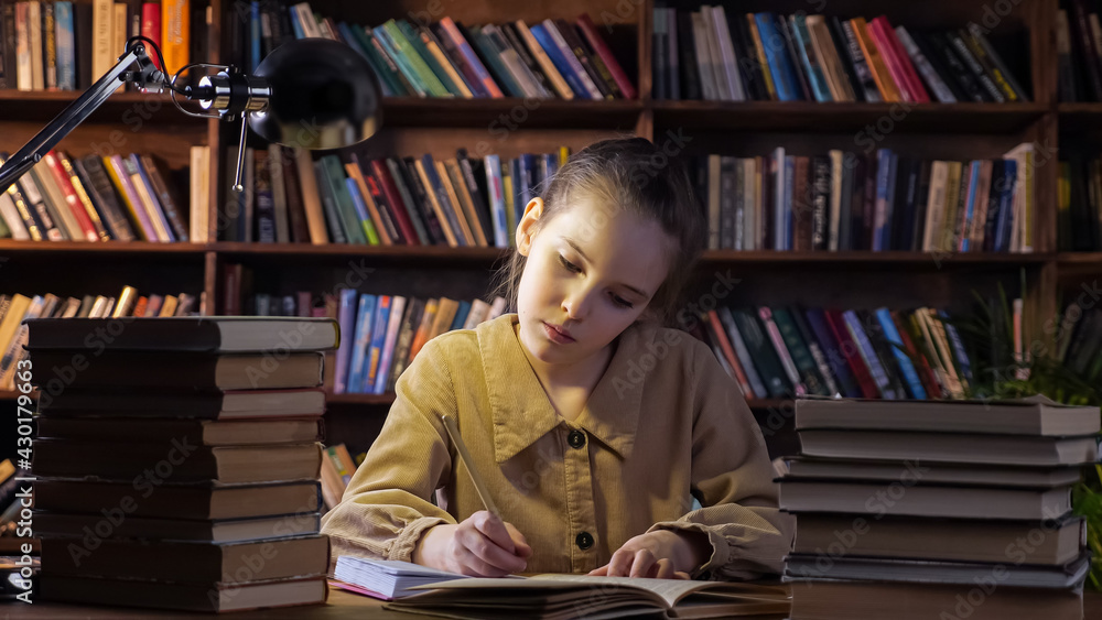 Concentrated girl in brown jacket writes homework in paper notebook with pencil sitting under lamp l