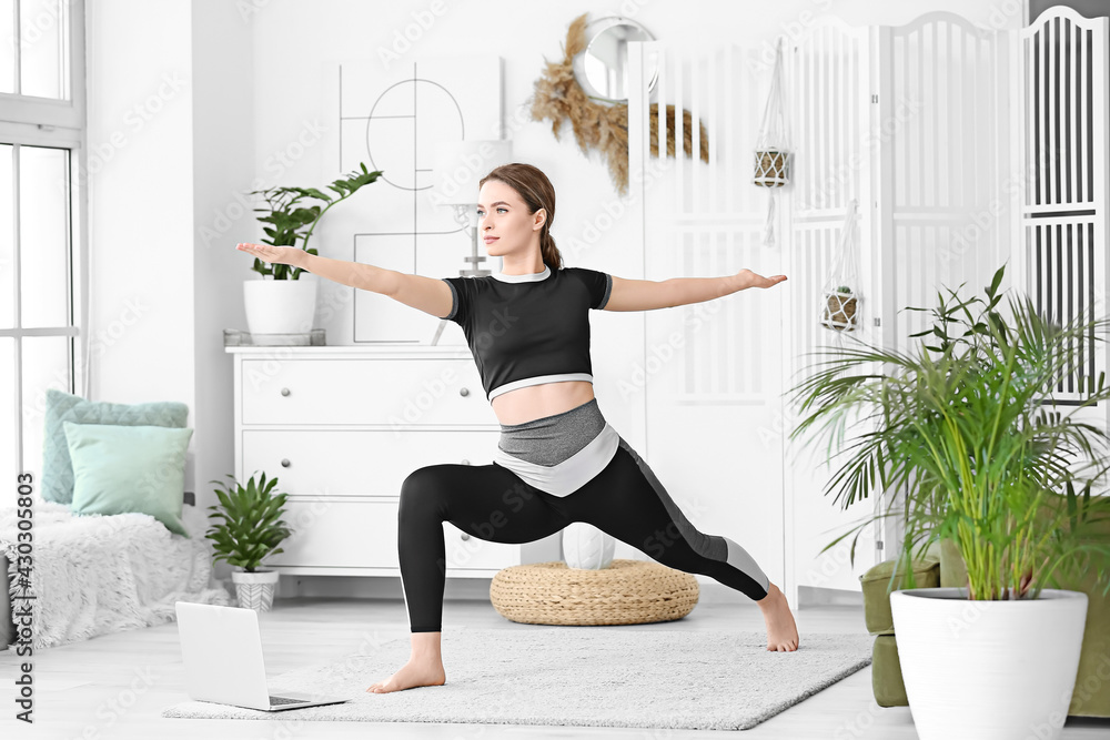 Sporty young woman with laptop practicing yoga at home during lockdown