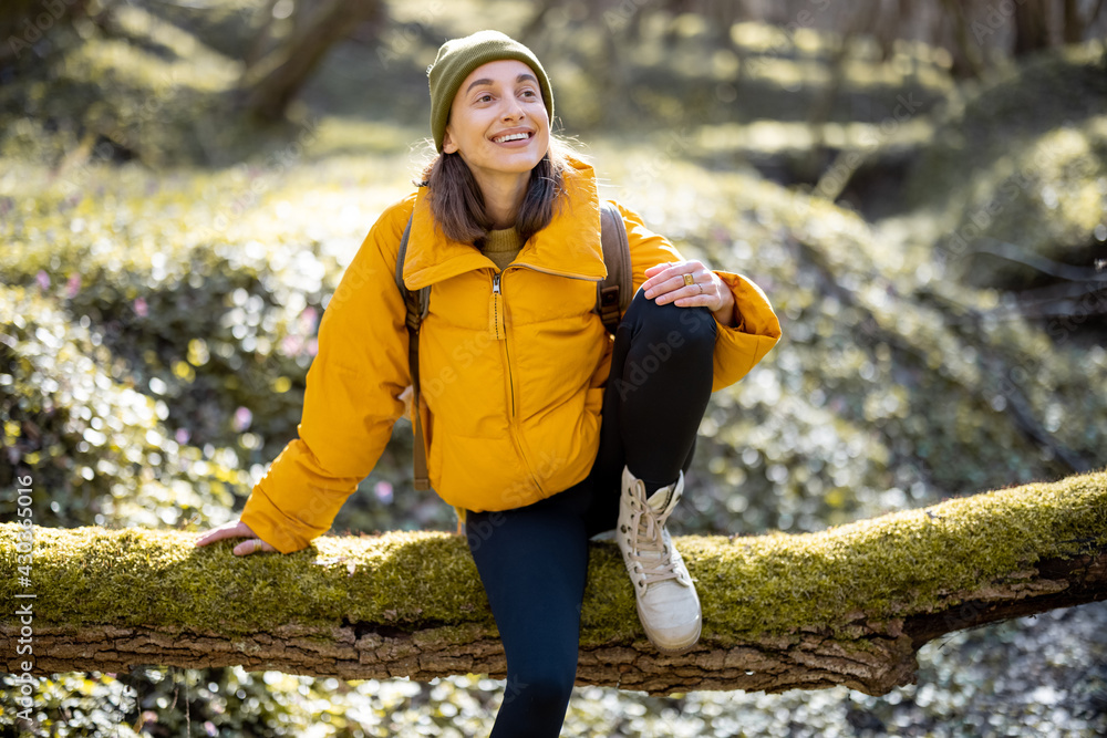 Young woman in hiking colorful clothes and backpack smiling and sitting on a tree above the river. E