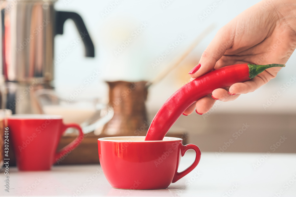 Woman dipping chili pepper into cup of coffee on table in kitchen