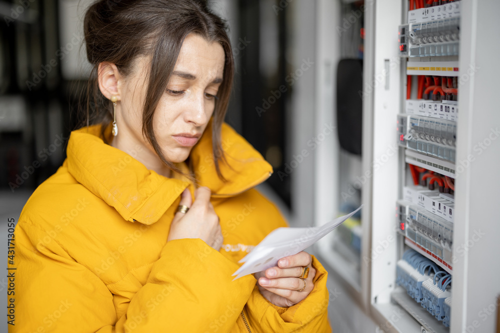 困惑的女人看着电费单，将数据与配电板上的电表进行比较。