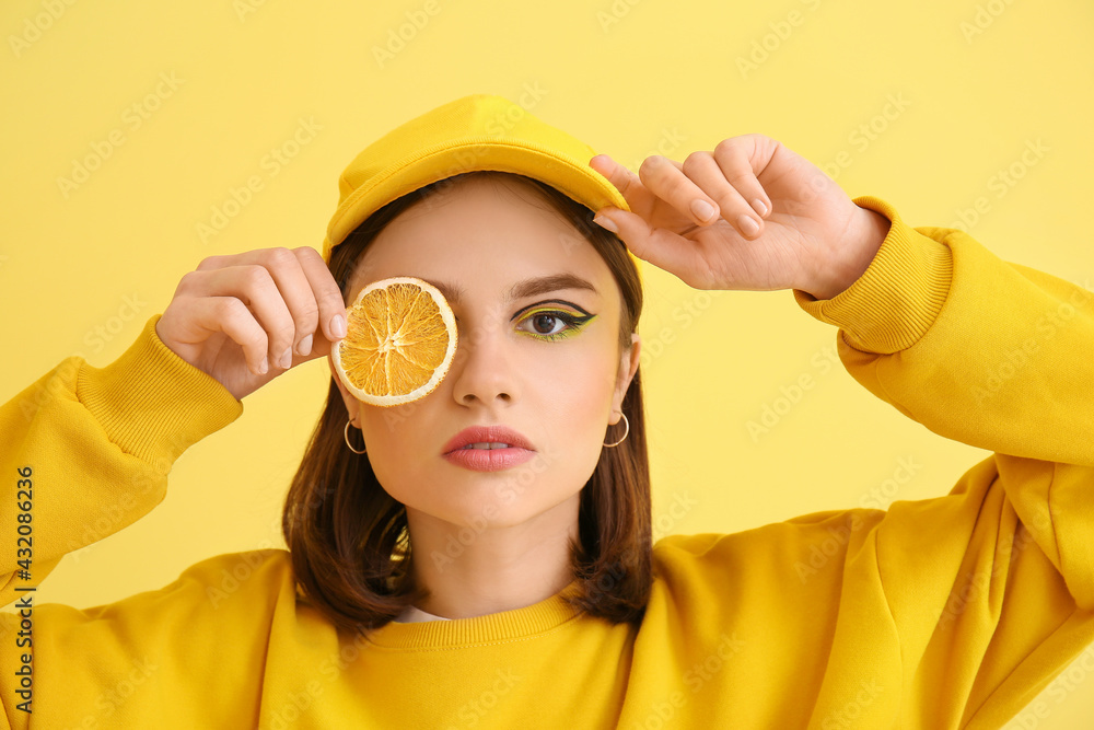 Stylish young woman with orange slice on yellow background