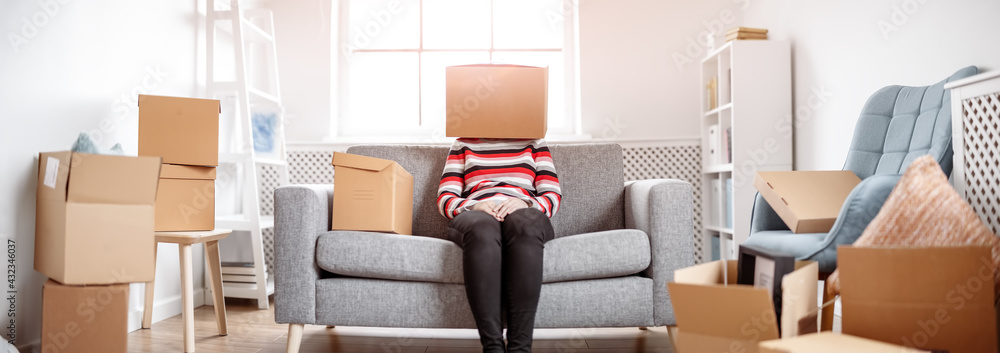 Tired womanwith cardboard box on her head sitting on the sofa in her new house.