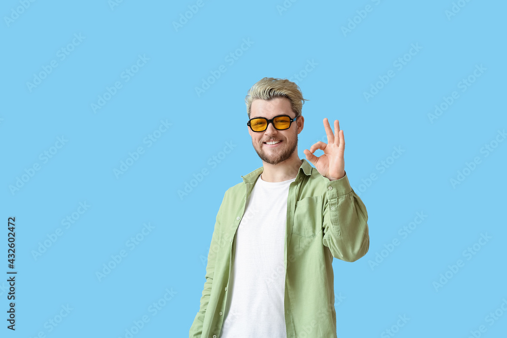 Young man with stylish sunglasses showing OK on color background