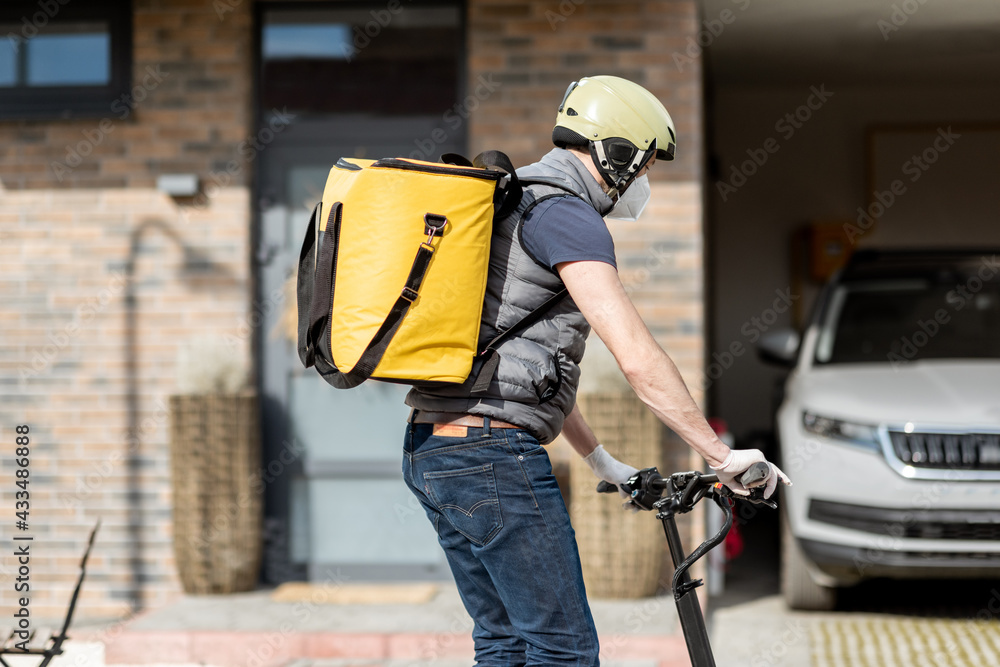 Delivery man in helmet with yellow backpack rides electro scooter on the street and looking for the 