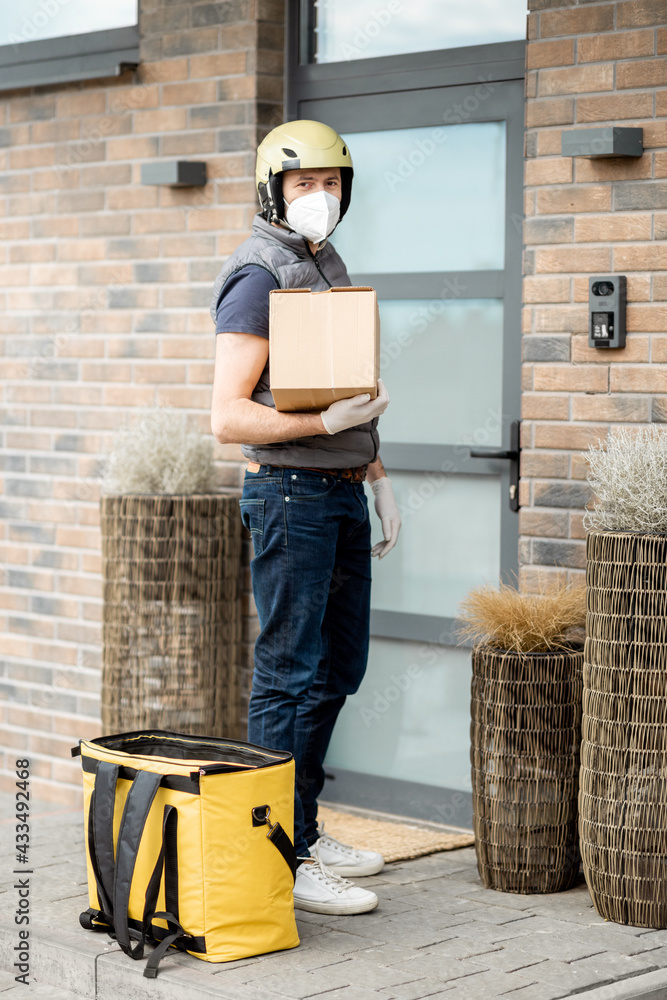 A portrait of courier in green helmet wearing mask and gloves holds a parcel box and staying near fr