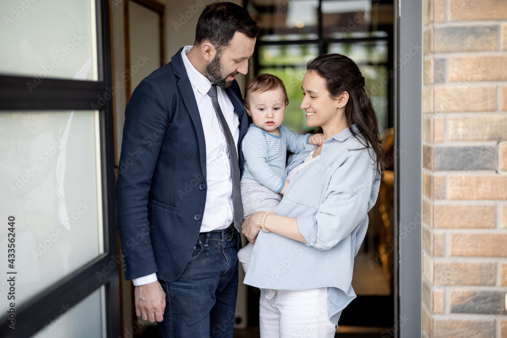 Happy husband hugs wife with one year old baby boy while standing in front of their front door house