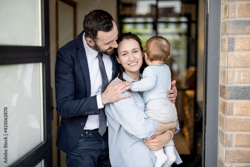 Happy husband hugs wife with one year old baby boy while standing in front of their front door house