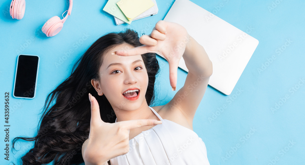 Young Asian girl lying on blue background with laptop, smartphone, headphones and note around