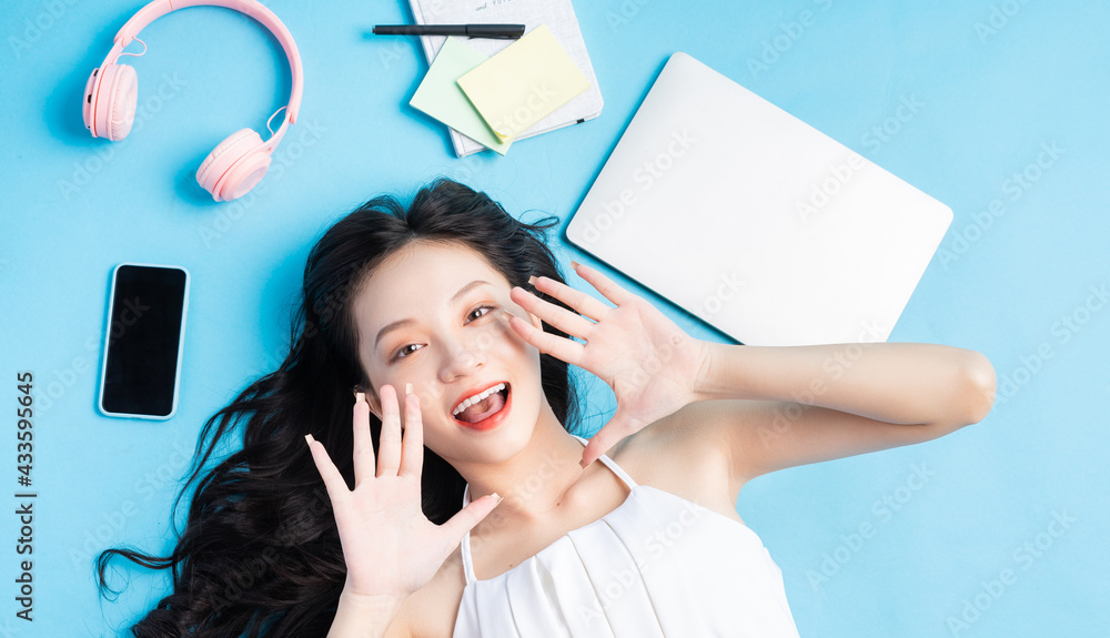 Young Asian girl lying on blue background with laptop, smartphone, headphones and note around