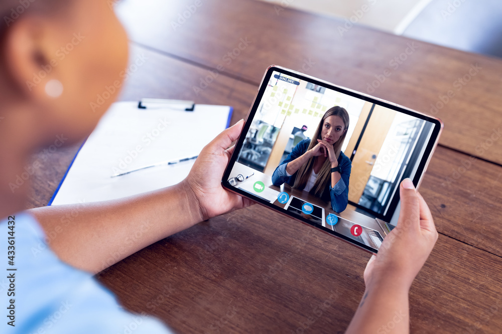 Mixed race businesswoman sitting at desk using tablet having video call with female colleague