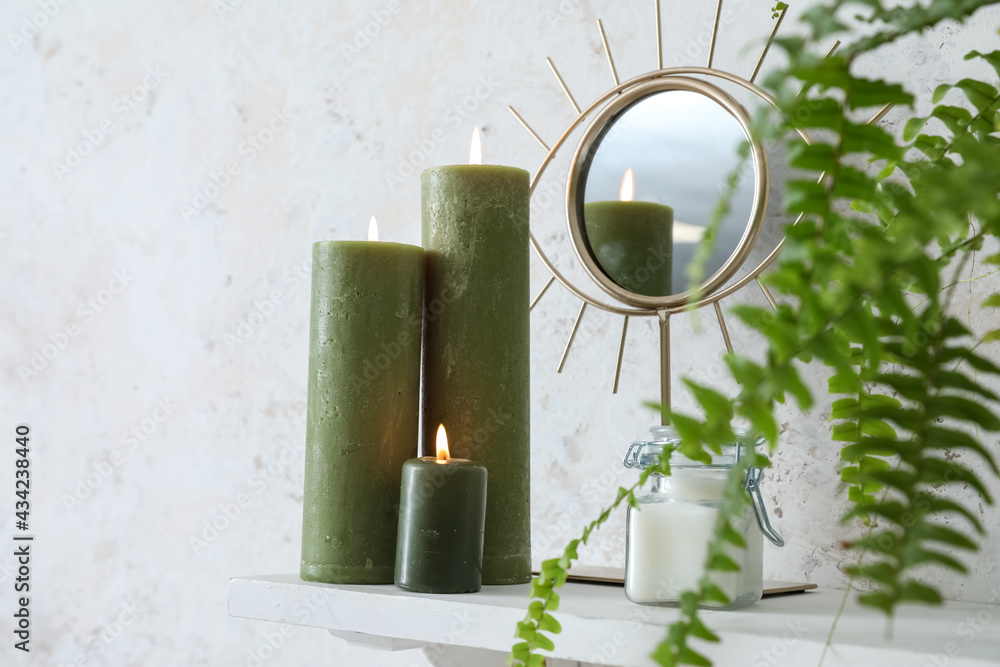 Burning candles, mirror and houseplant on shelf near light wall