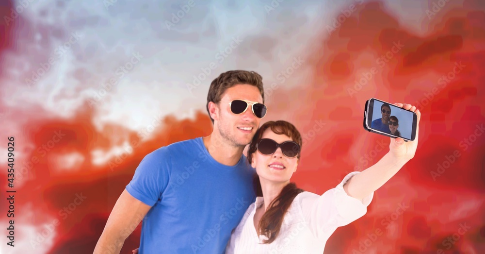 Composition of happy holiday couple in sunglasses smiling, taking selfie , over sunset sky