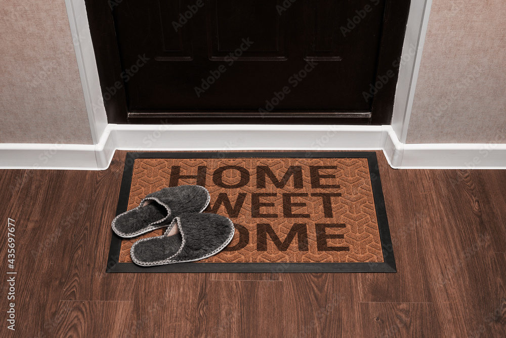 Doormat with text Home sweet home before the dark door in the hall. Home slippers on the rug. Top vi