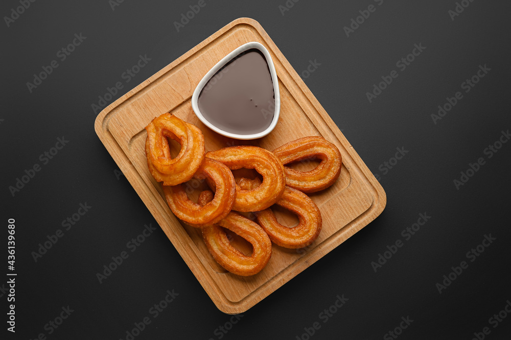 Board with tasty churros and melted chocolate sauce on dark background