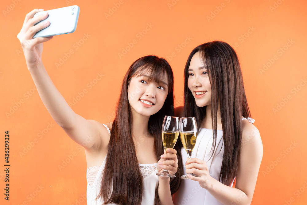 Two beautiful young Asian girls raising wine glasses on orange background