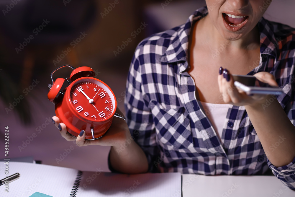 Angry woman with alarm clock talking by mobile phone in office at night