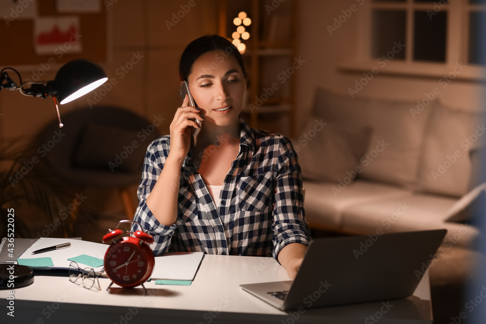 Beautiful woman talking by mobile phone while using laptop in office at night