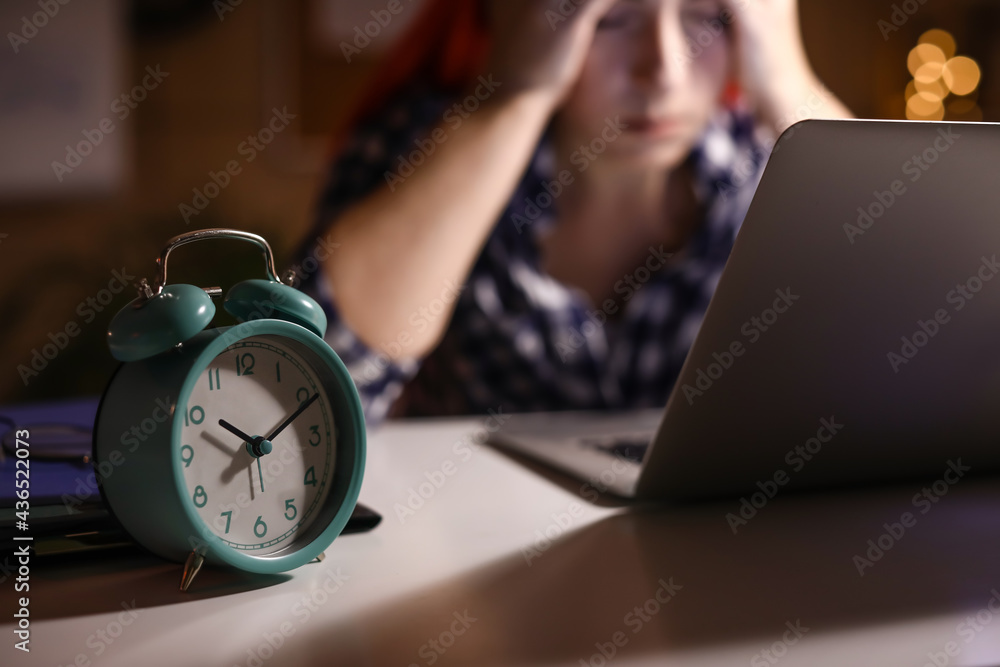 Alarm clock on table of tired woman working with laptop in evening