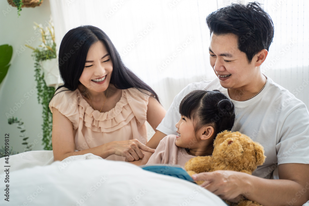 Asian family parent read storytale book to young daughter in bedroom.