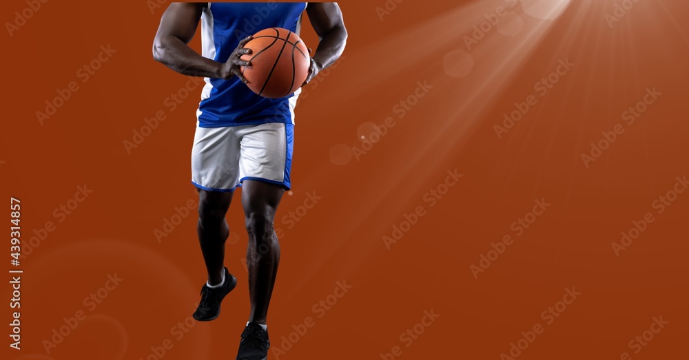 Mid section of male basketball player holding basketball against spot of light on orange background